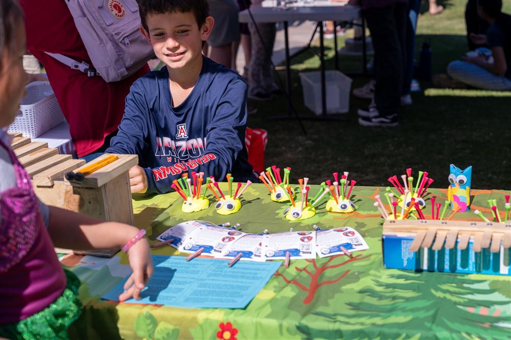 A student smiles by his booth filled with hand made musical instruments and crafts that look like cardboard bugs.